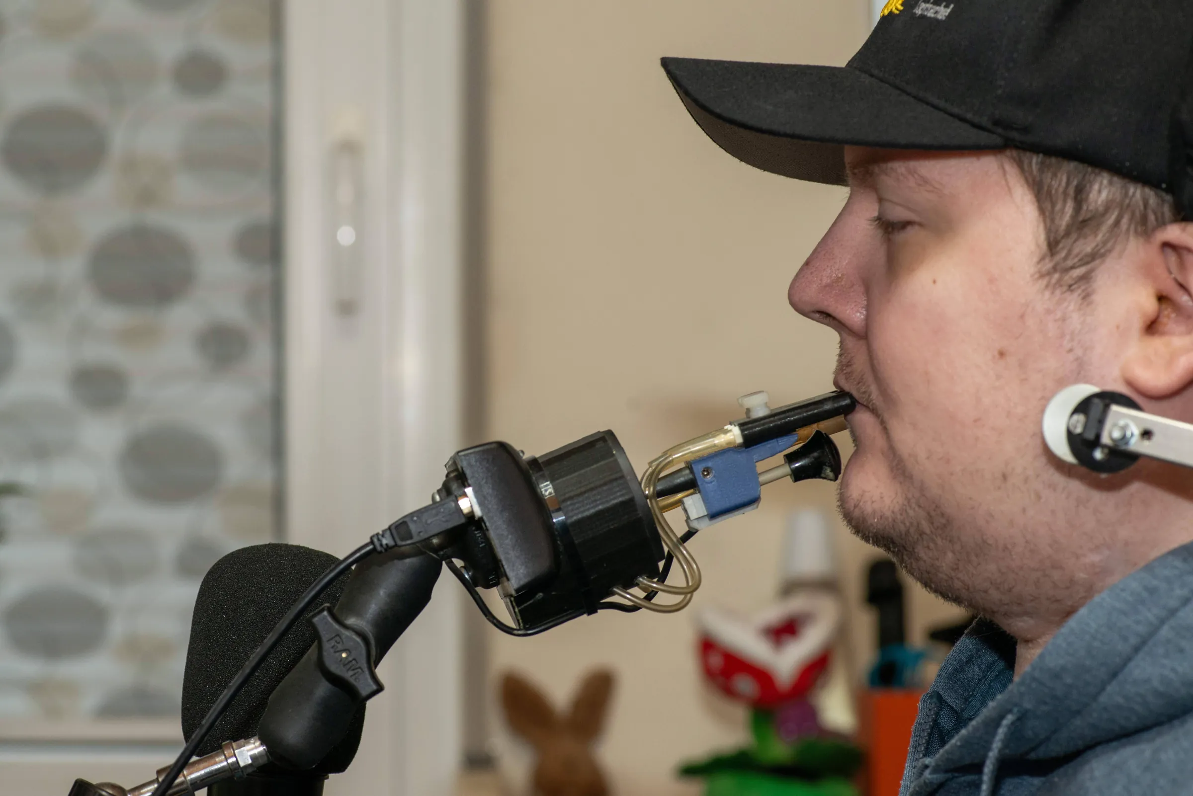 A man in a cap uses a QuadStick. The mouthpiece is at his lips and the device sits on a mounting arm in front of him.
